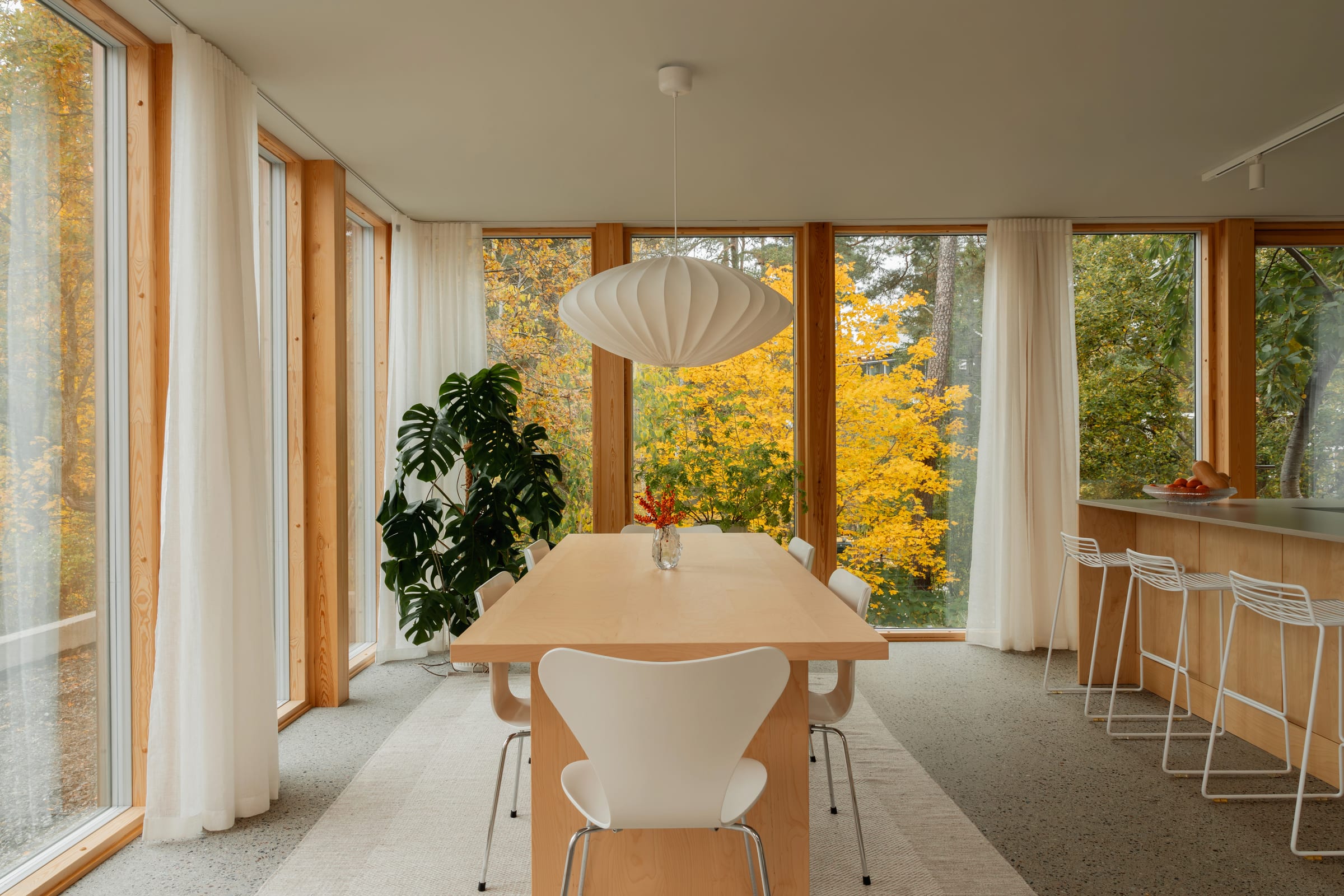 Matplats med långbord i björk och vita stolar framför stora fönster, med köket till höger